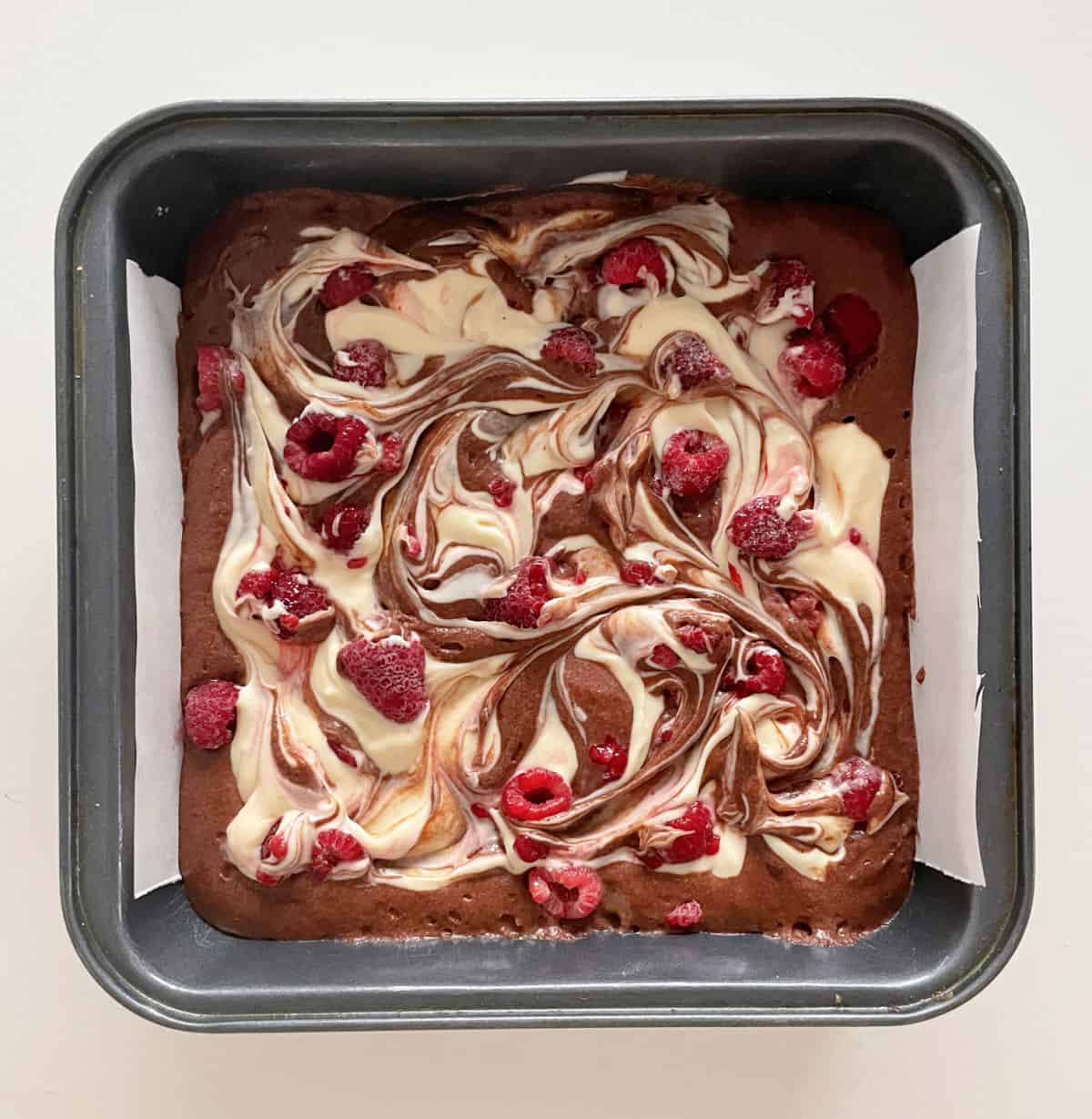 Cheesecake swirl brownie batter with raspberries ready to be baked in a metal square pan. 