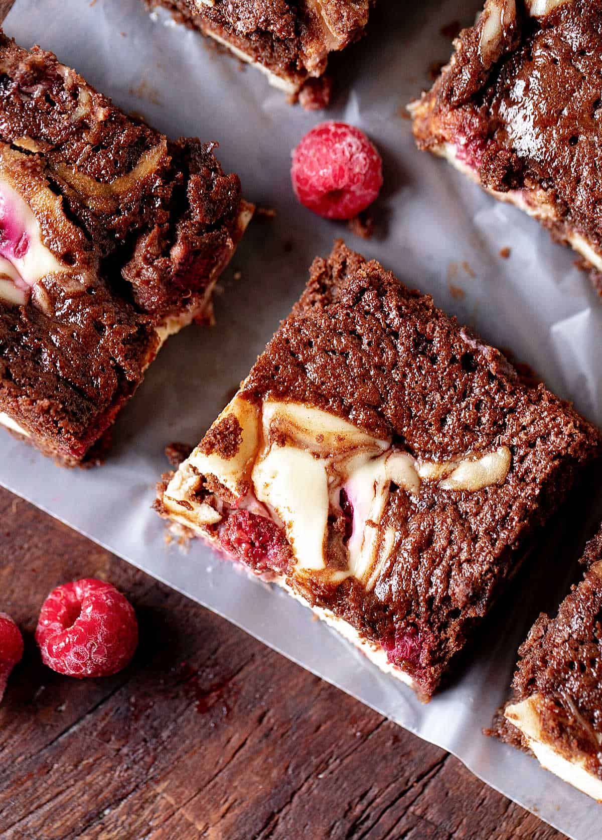 Close up overview of cheesecake brownie squares with raspberries on a plastic sheet and wooden board.
