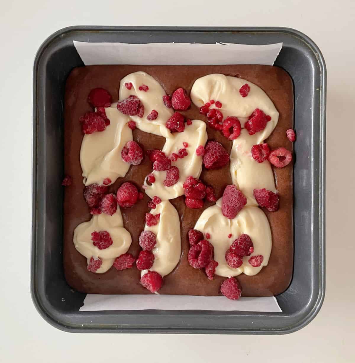 Layered brownie batter, cheesecake mix, and raspberries in a square metal pan on a white surface.