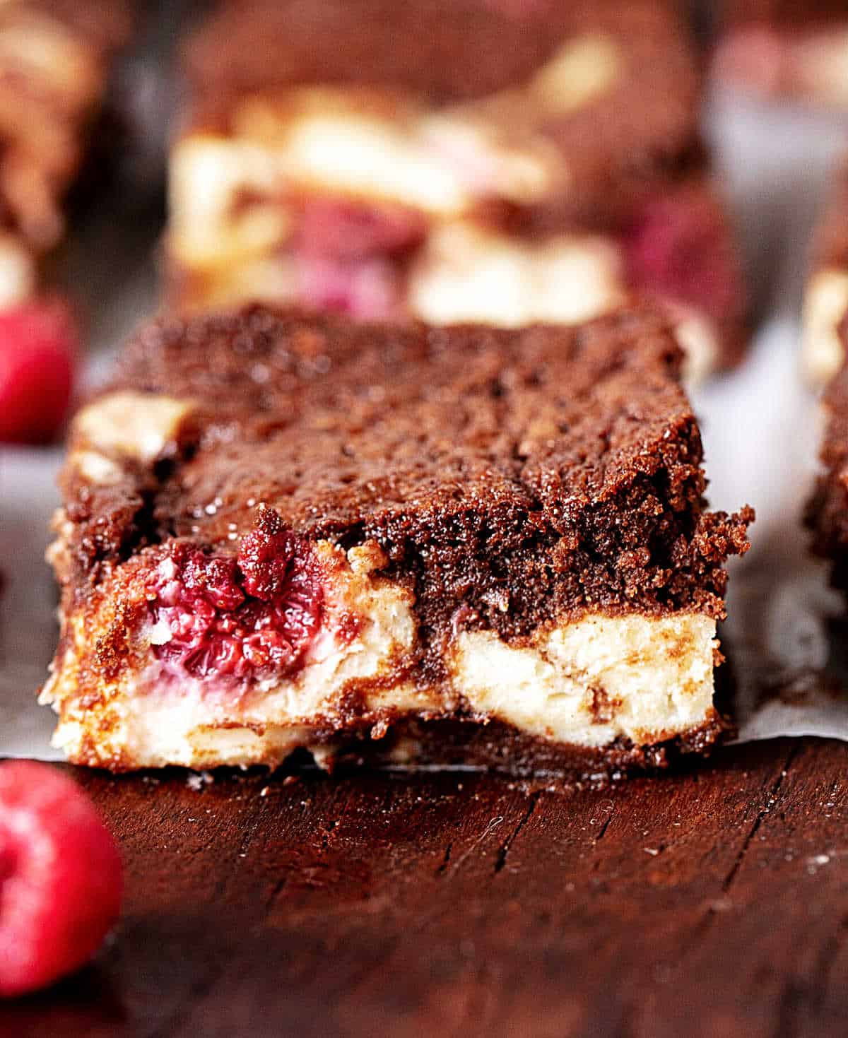 Close up of brownies with cheesecake swirl and raspberries on a dark wooden board.