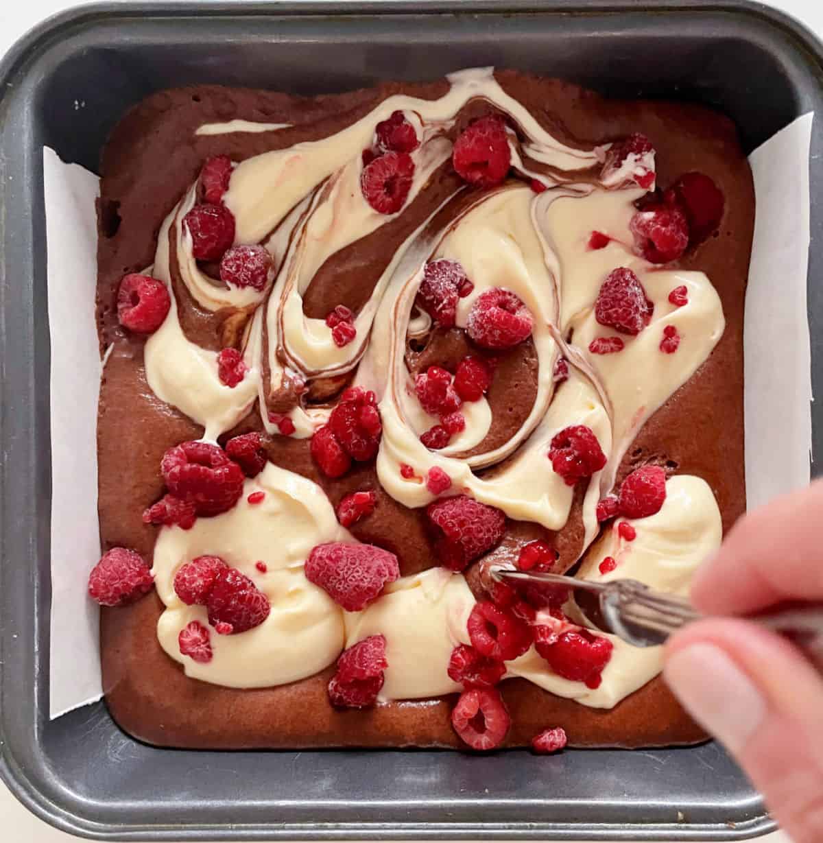 Swirling cheesecake and brownie batters with raspberries with a knife. Metal square pan. 