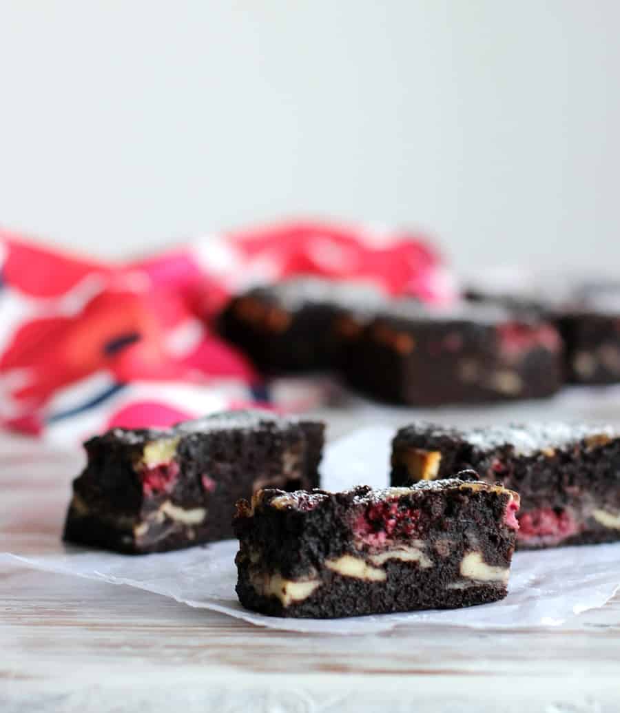 Several rectangles of cheesecake brownies with rapsberries on white surface, colorful blurred background