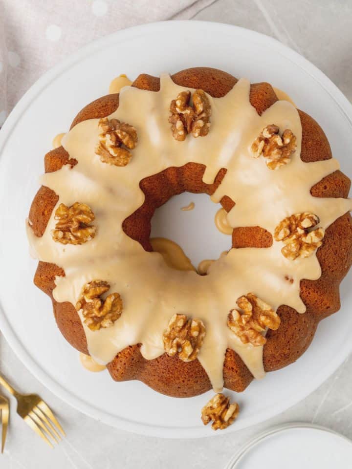 Top view of whole glazed walnut bundt cake on a white plate, gold forks, and light-colored cloth.
