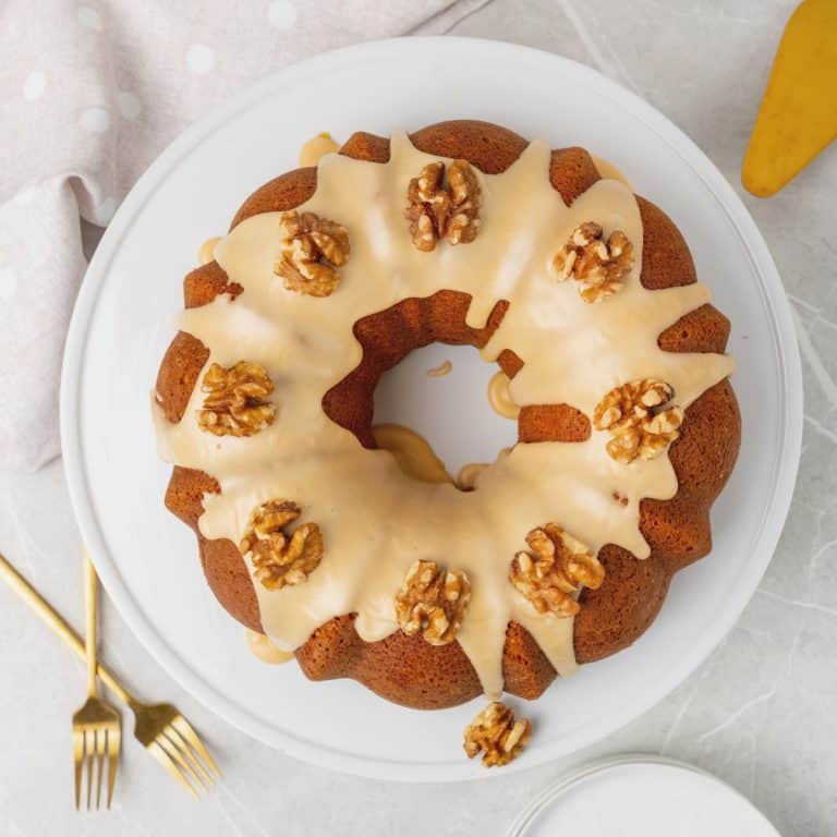 Top view of whole glazed walnut bundt cake on a white plate, gold forks, and light-colored cloth.