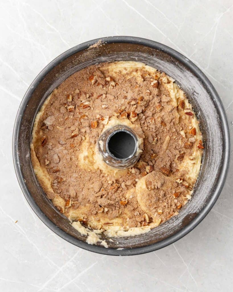 Cinnamon pecan mixture sprinkled on cake batter in a tube pan on a light grey surface.
