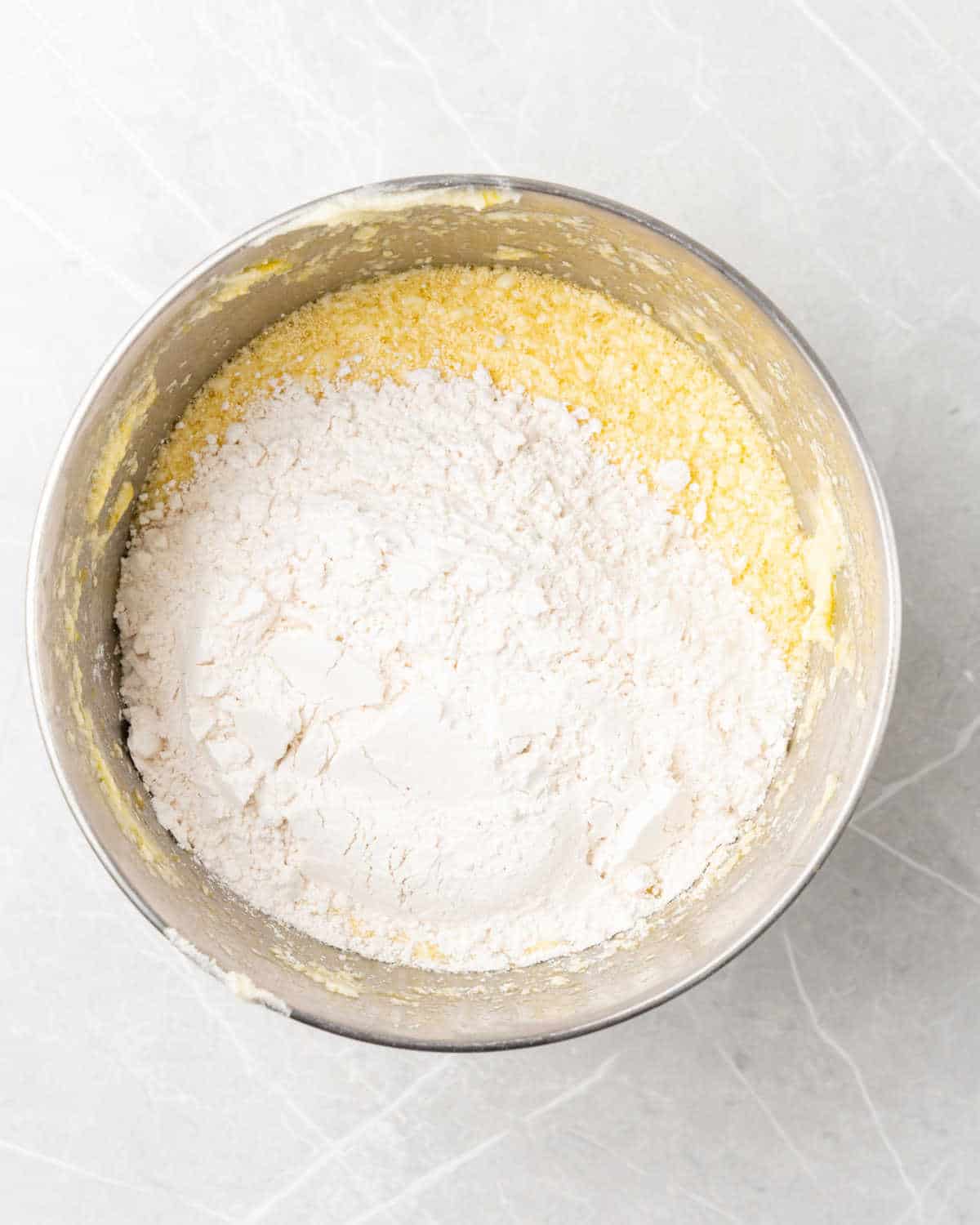Flour added to cake batter in a metal mixer bowl on a grey surface. 