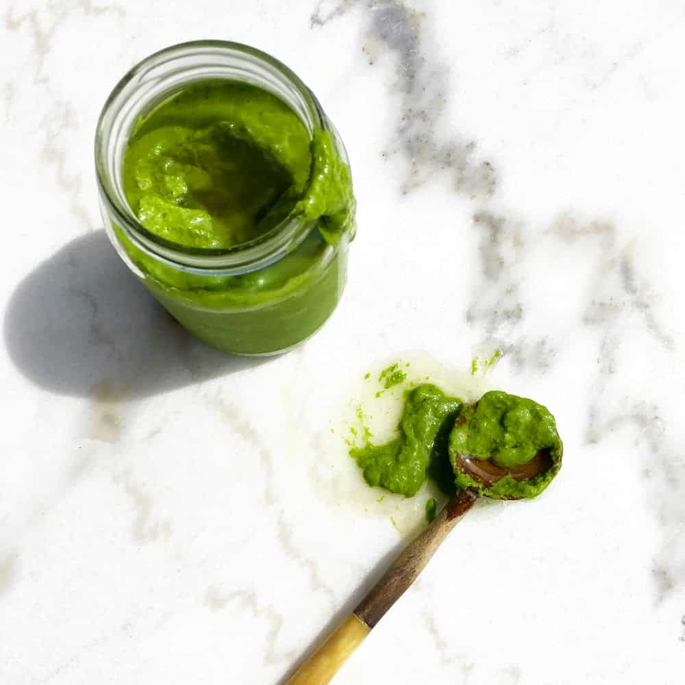 Glass jar and wooden spoon with salsa verde on marble surface.