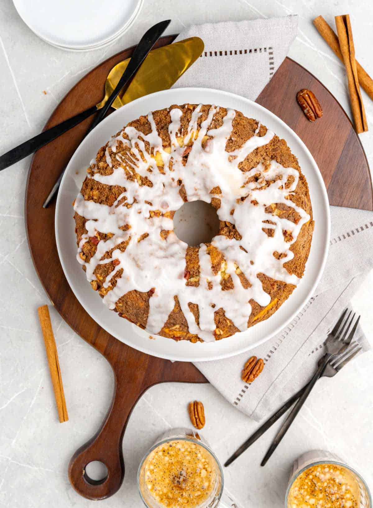 Whole glazed coffee cake on a white plate set on a round wooden board. Grey surface with coffee cups and forks.