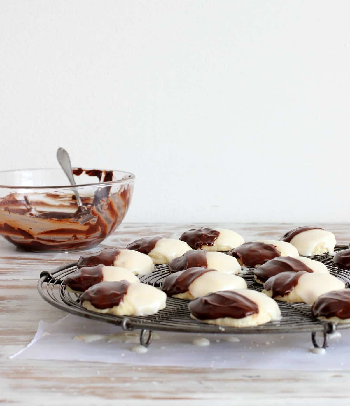 Glazed black and white cookies on a cooling rack with a bowl of ganache in the white background. 