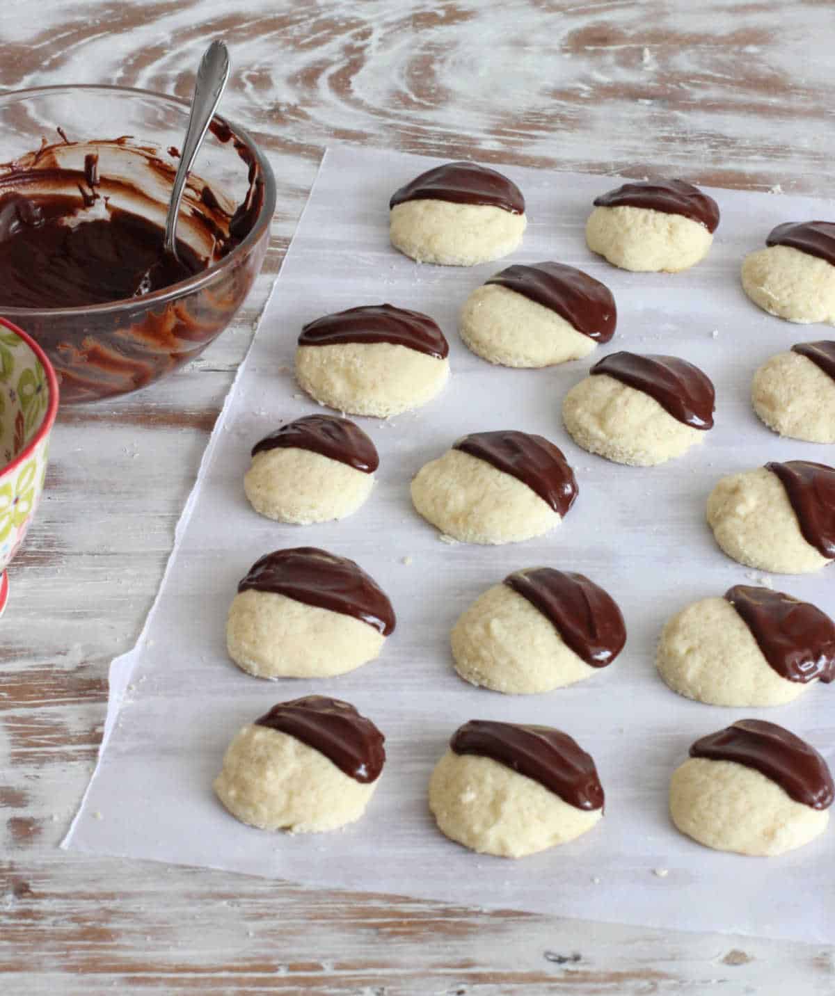 Half glazed vanilla cookies on parchment paper on a streaked white table. A bowl containing chocolate icing.
