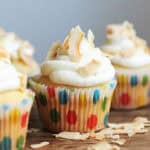 One central white frosted cupcake in dotted paper liner on wooden table with coconut flakes.