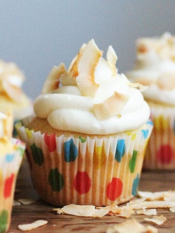 One central white frosted cupcake in dotted paper liner on wooden table with coconut flakes.