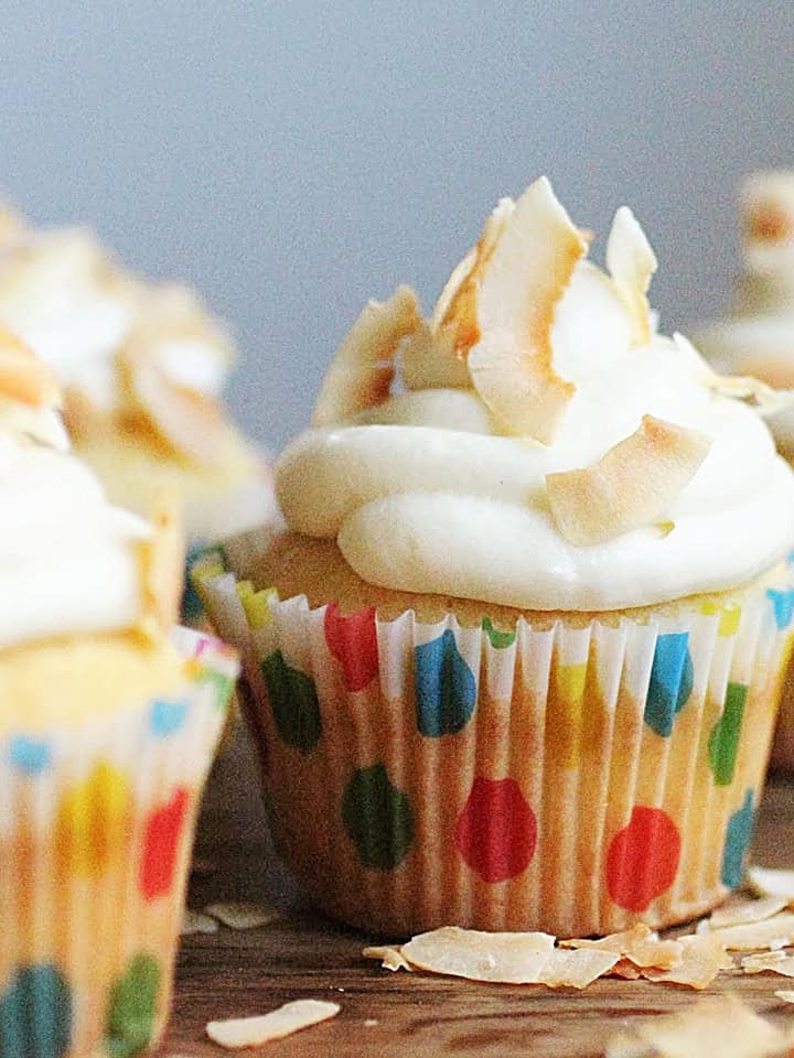 One central white frosted cupcake in dotted paper liner on wooden table with coconut flakes.