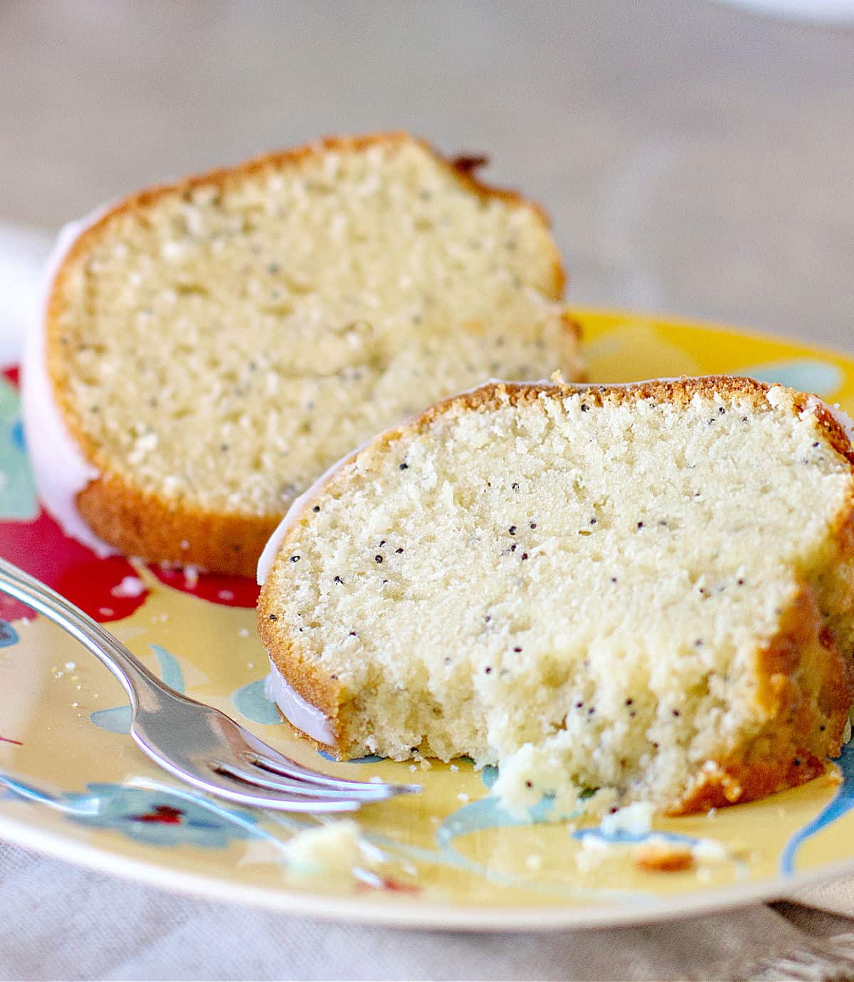 One partially eaten slice of poppy seed bundt cake with whole one on a colorful plate, a silver fork.