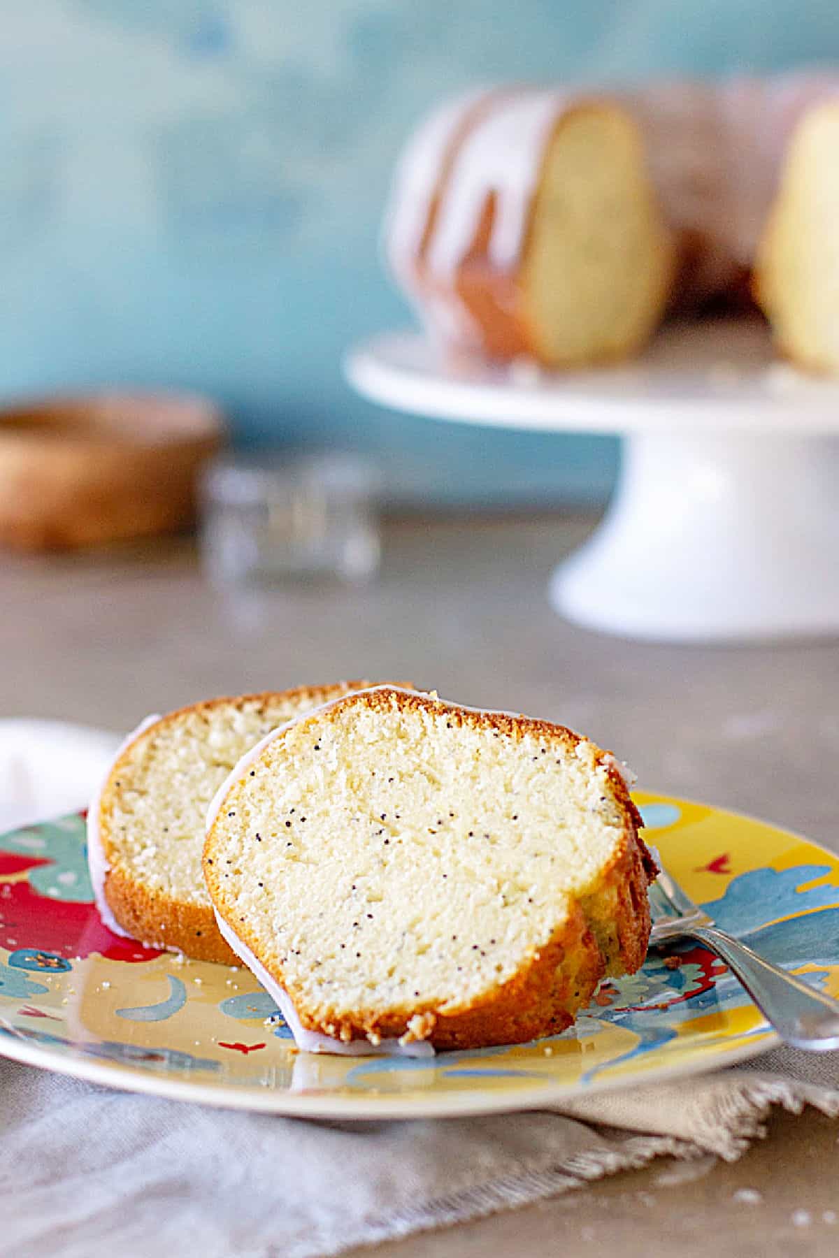 Glazed slices of poppy seed cake on a colorful plate, blue background. Whole cake on a white stand.