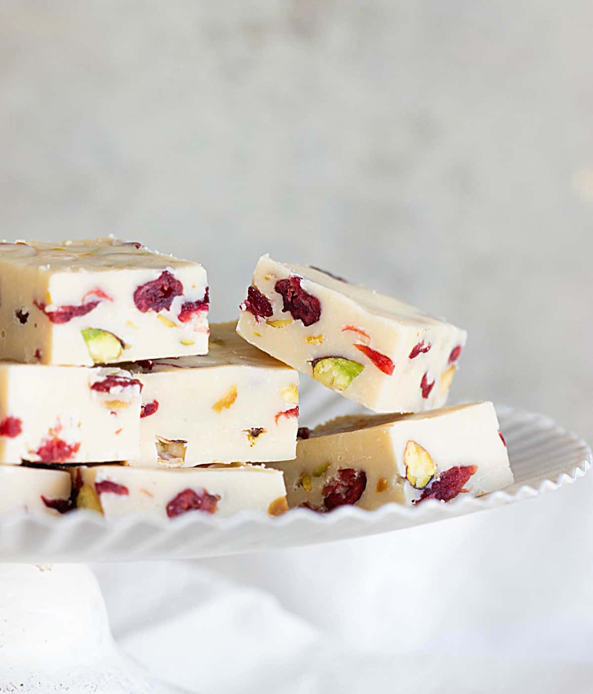 Squares of white fudge with cranberries and pistachios on a white cake stand, light grey background
