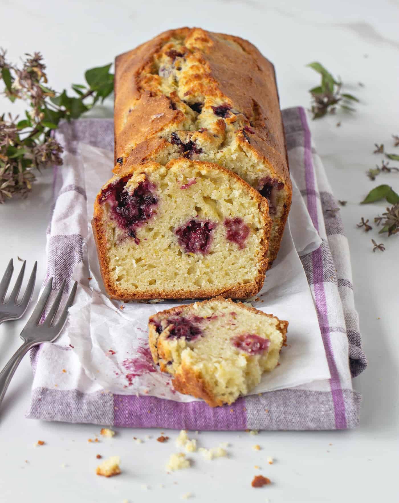 Loaf of sliced Orange blackberry cake, kitchen towel, forks, white surface, small flowers