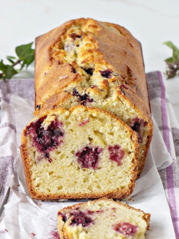 Muffin loaf with blackberries whole and sliced on white purple cloth. White background, green leaves around.