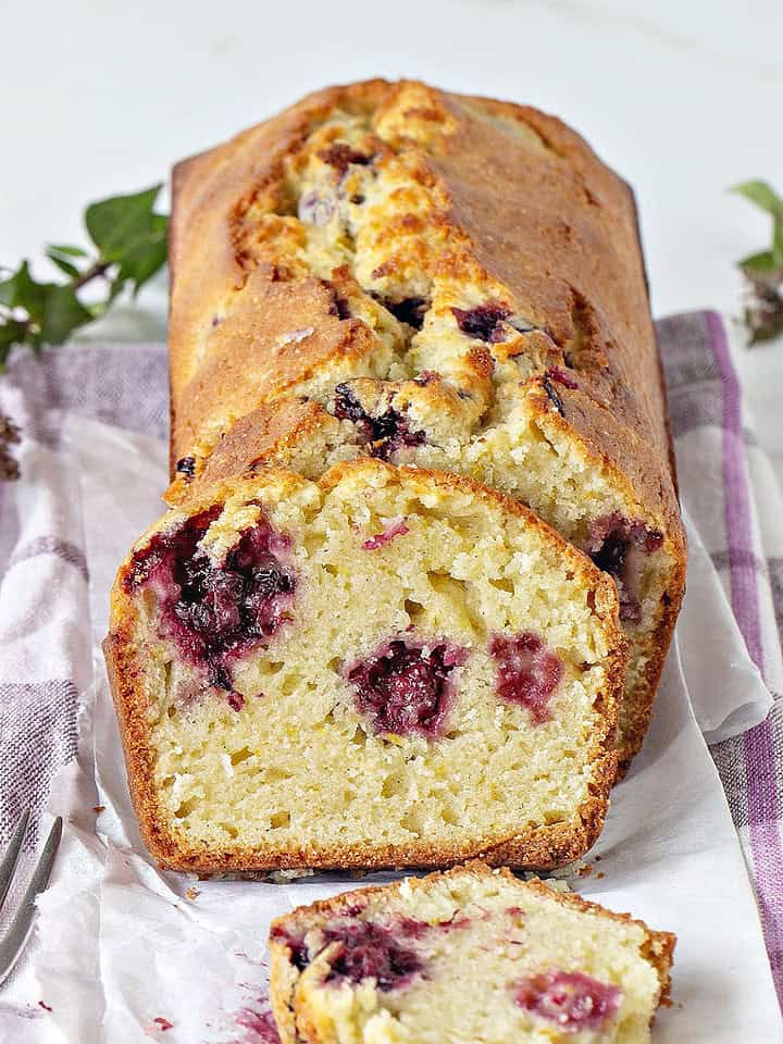 Muffin loaf with blackberries whole and sliced on white purple cloth. White background, green leaves around.