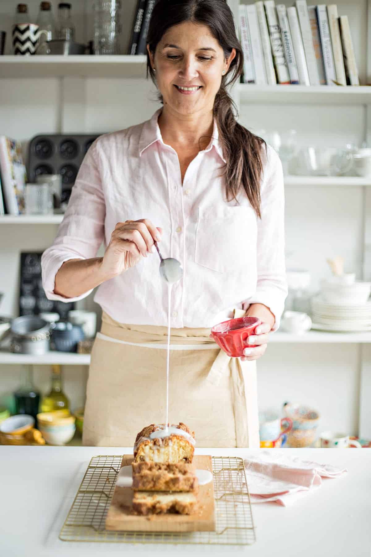 Woman with pink shirt and brown apron glazing loaf coffee cake, prop library as background
