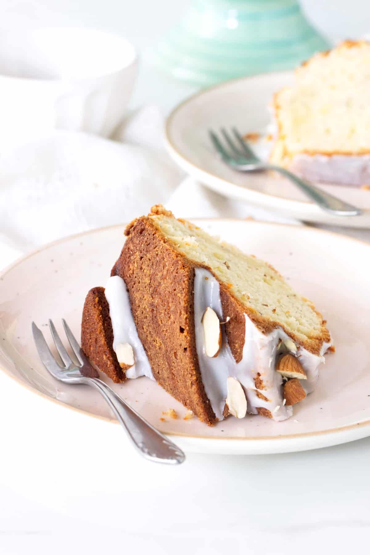 Slice of glazed almond bundt cake on a plate with fork, another slice in background