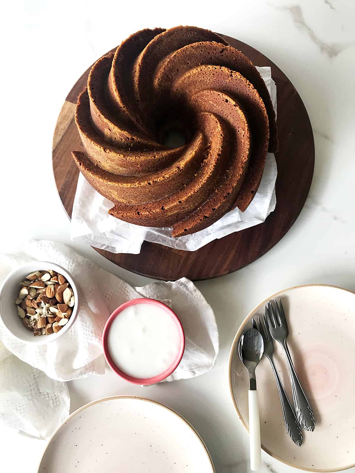 Bundt cake on wooden round board, plates, forks and bowls with almonds and glaze