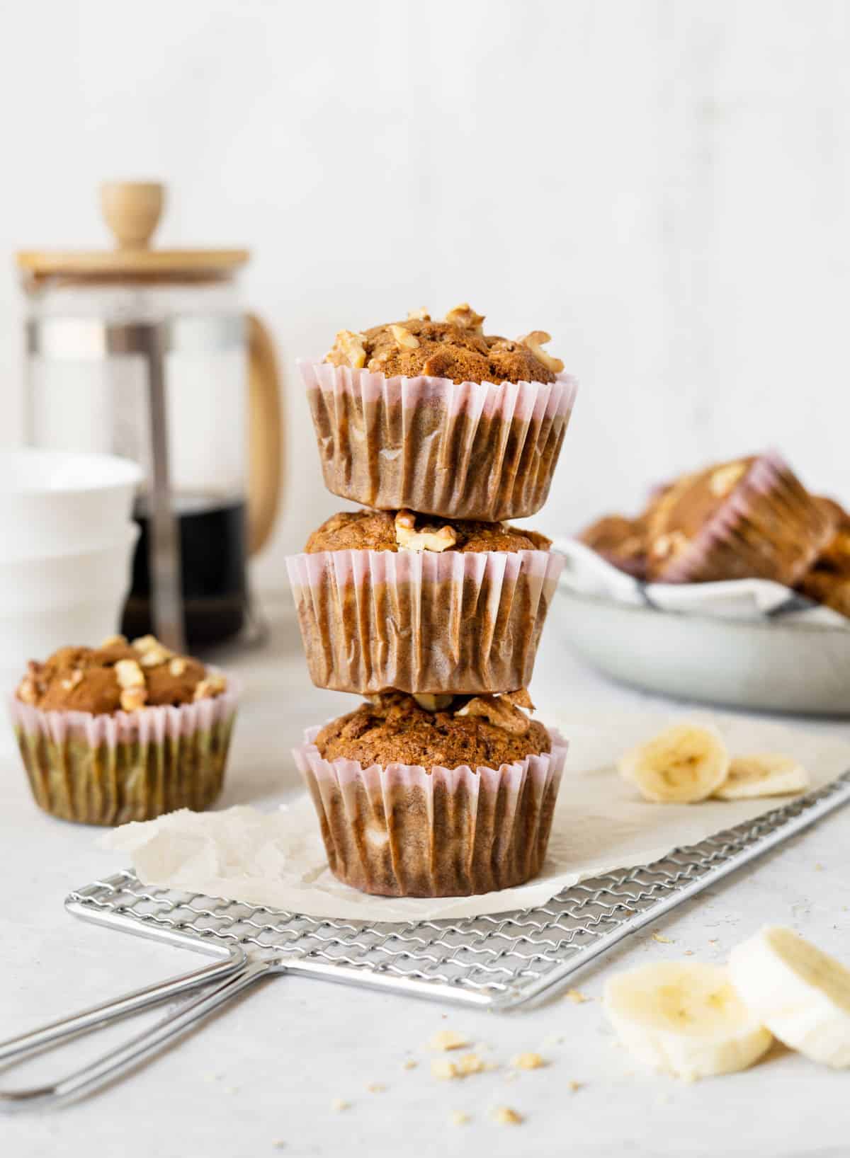 Stack of three banana muffins in paper liners on parchment paper and a rack. White surface and background with coffee maker and more muffins. 