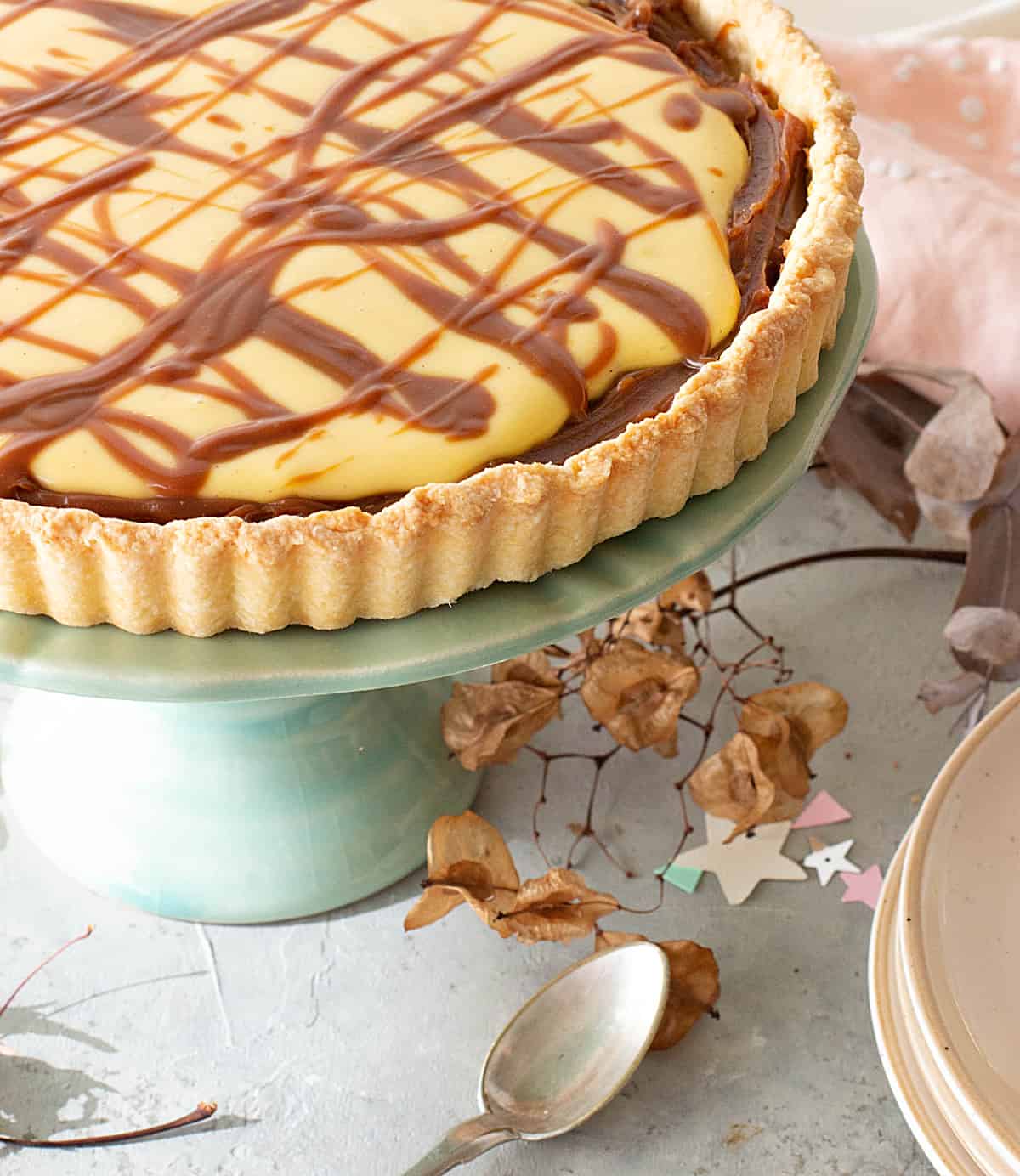 Close up of lemon tart with dulce de leche on green cake stand, grey surface with dried leaves, spoon, stack of plates.