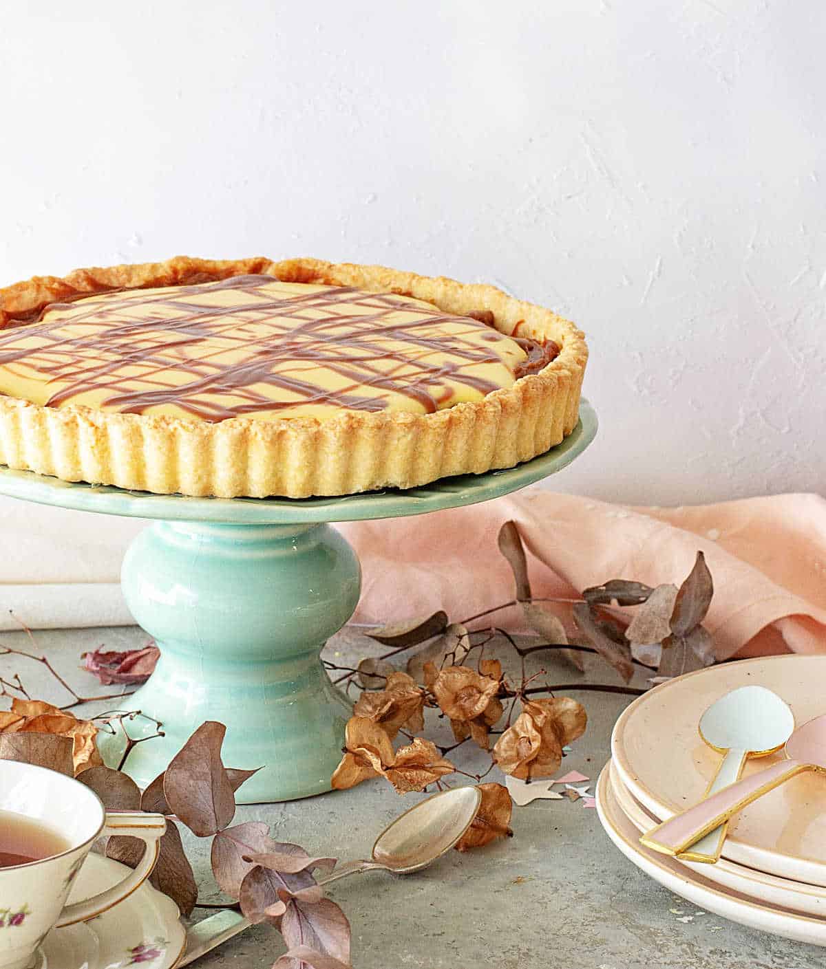 Whole lemon tart on a green cake stand. Stack of pink plates, dried leaves, a grey surface. 