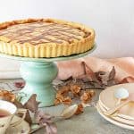 A lemon tart on a green cake stand. Stack of pink plates, dried leaves, a grey surface.