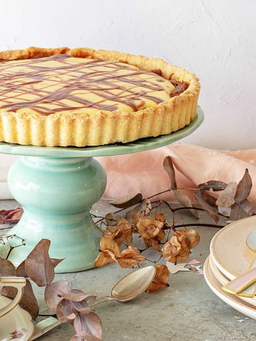 A lemon tart on a green cake stand. Stack of pink plates, dried leaves, a grey surface.