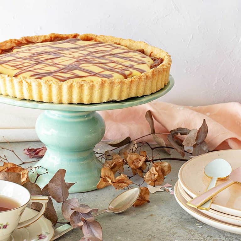 A lemon tart on a green cake stand. Stack of pink plates, dried leaves, a grey surface.