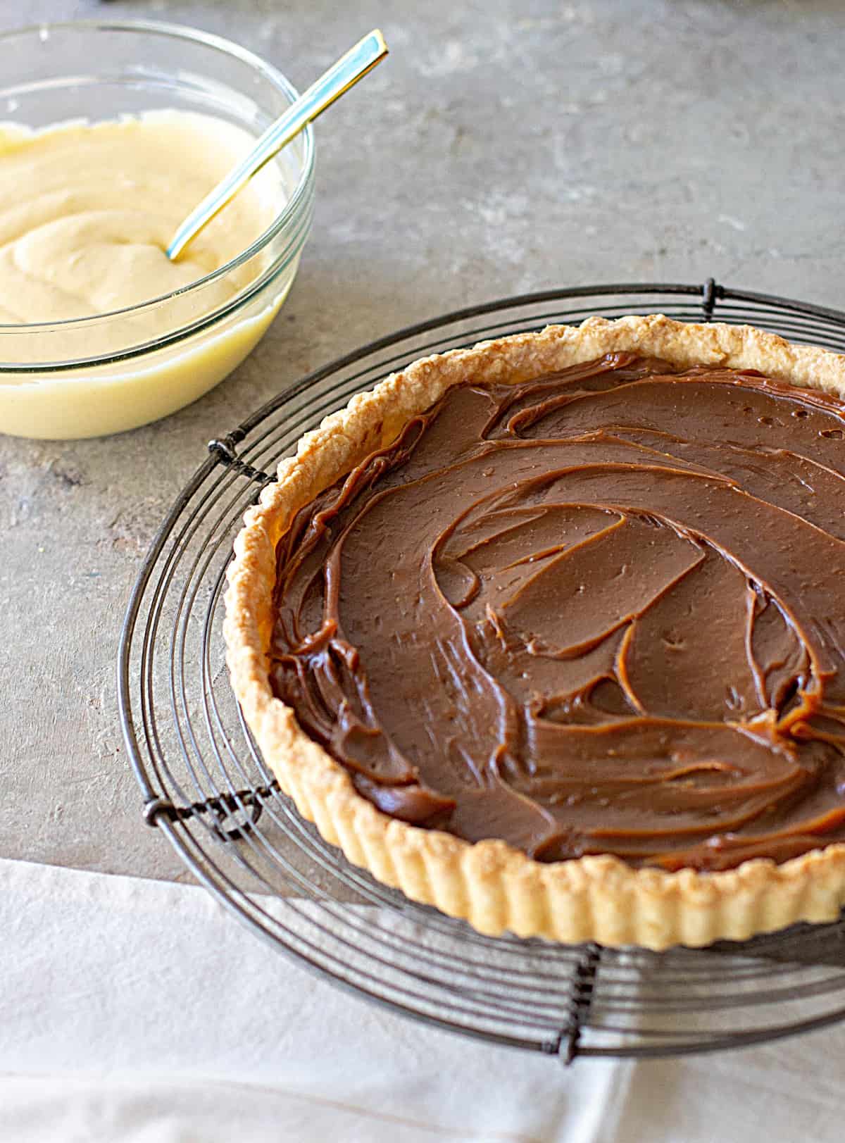 On a grey surface a wire rack with a pie tart dough with dulce de leche and bowl with lemon cream. 