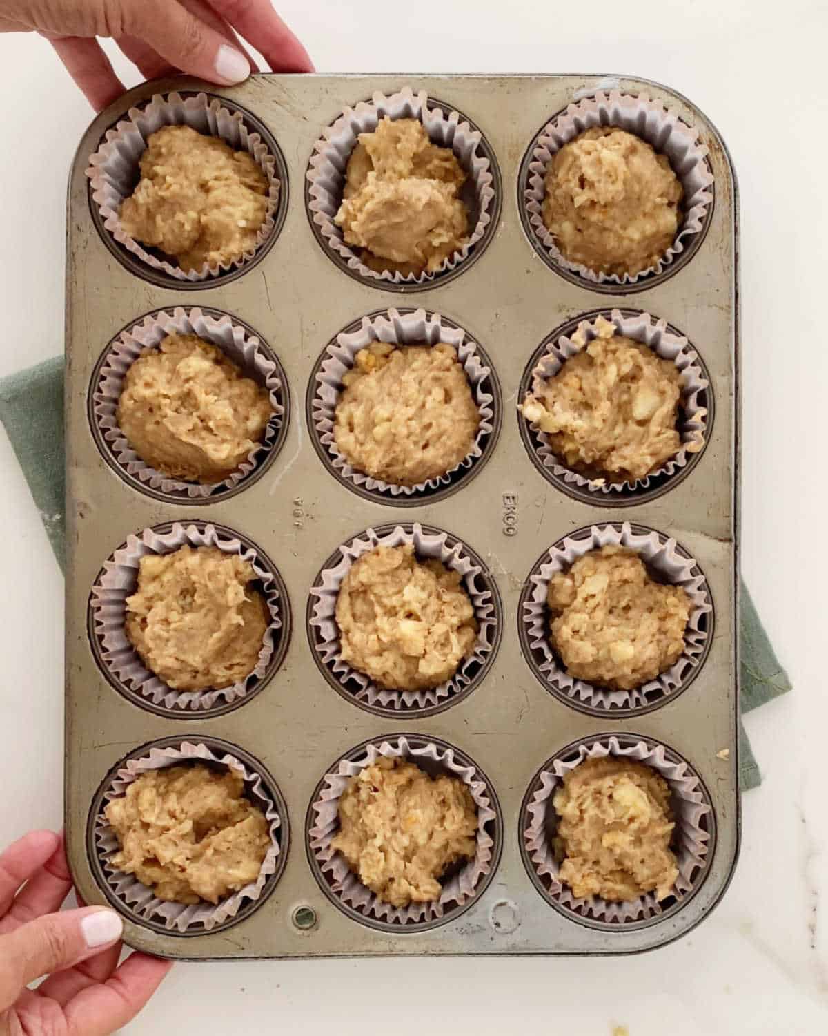 Metal muffin pan with paper liners filled with banana muffin batter. Hands holding it. White marble surface.