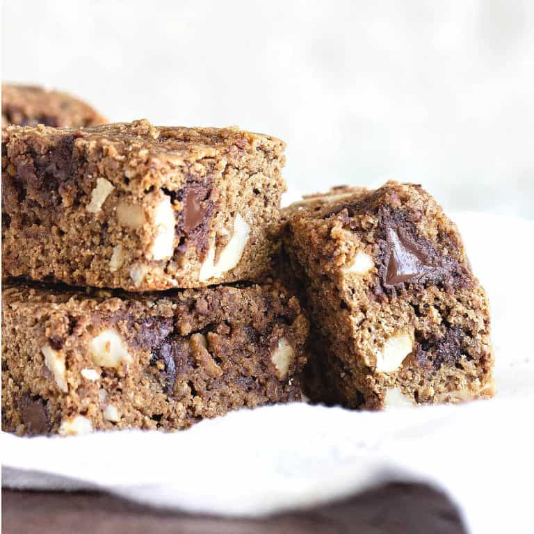 Squares of cookie blondies with chocolate chunks on a white cloth and white background.