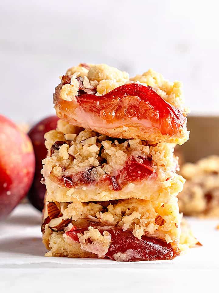 Stack of plum crumble bars. Whitish surface and background. Whole plums.