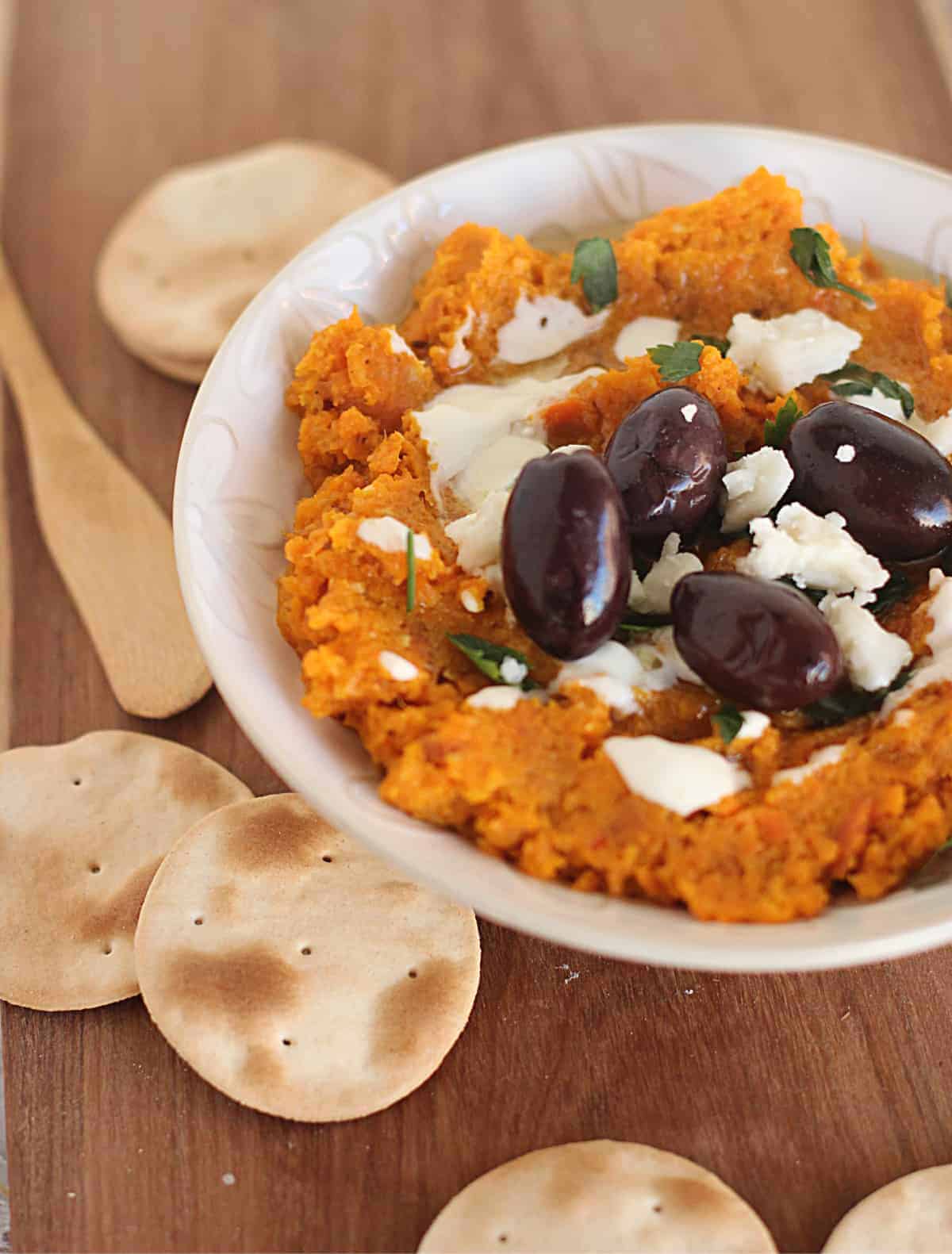 White Bowl of carrot dip, black olives, goat cheese and crackers on wooden board