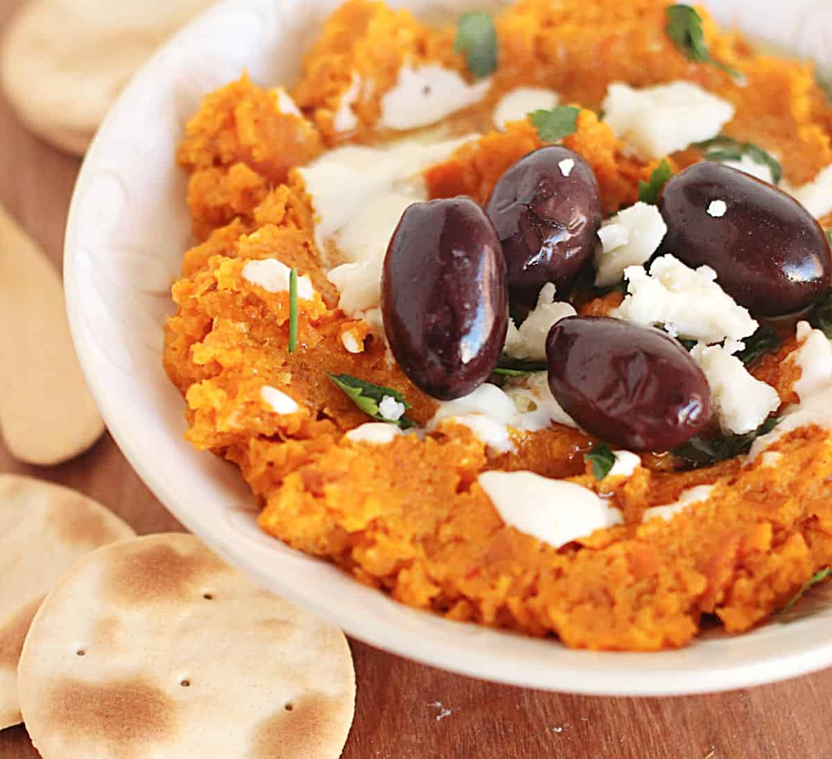 White bowl with carrot dip, black olives, goat cheese and crackers.