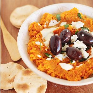 Creamy carrot dip with black olives and feta cheese in a white bowl with crackers.