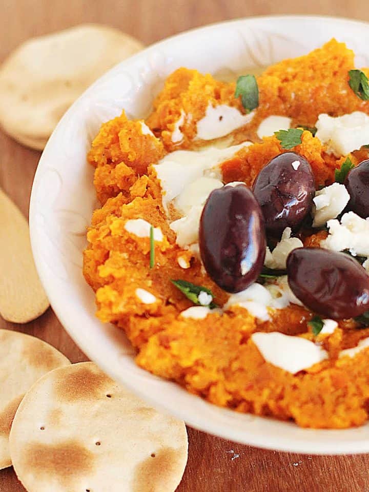 Creamy carrot dip with black olives and feta cheese in a white bowl with crackers.