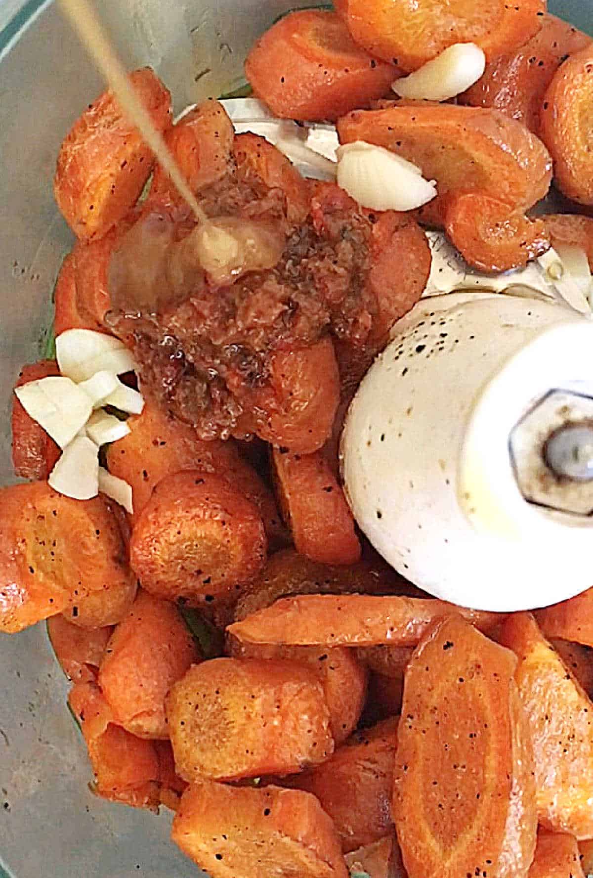 Carrots and spices in the bowl of a food processor with honey being poured in.