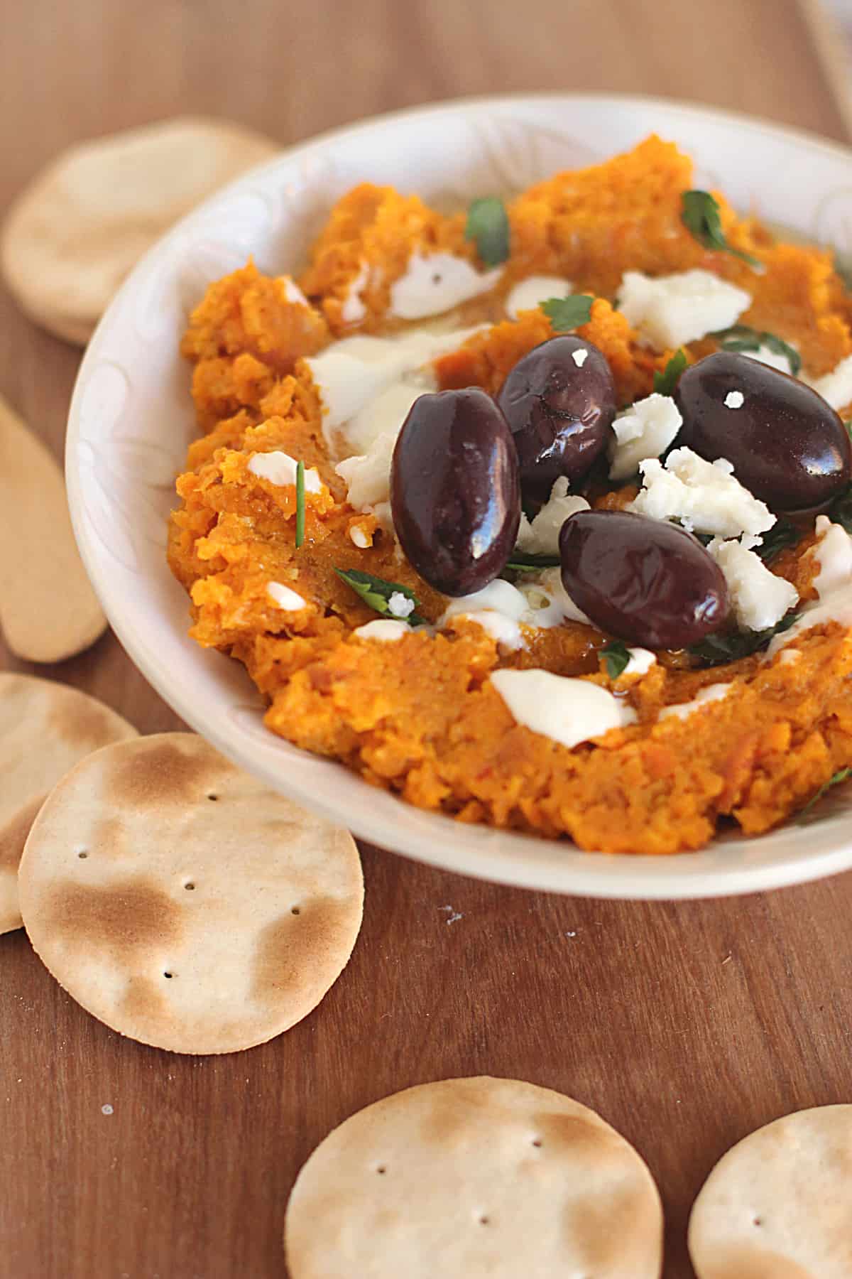 On a wooden board a white bowl with carrot dip and black olives surrounded by crackers