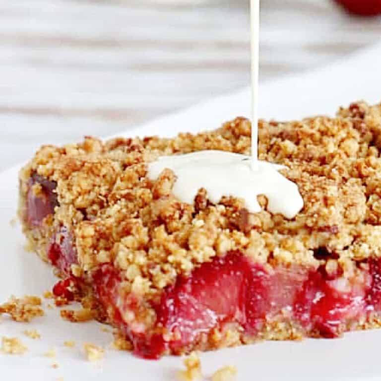 Slice of plum crumb bars on white plate, liquid cream being poured on top.