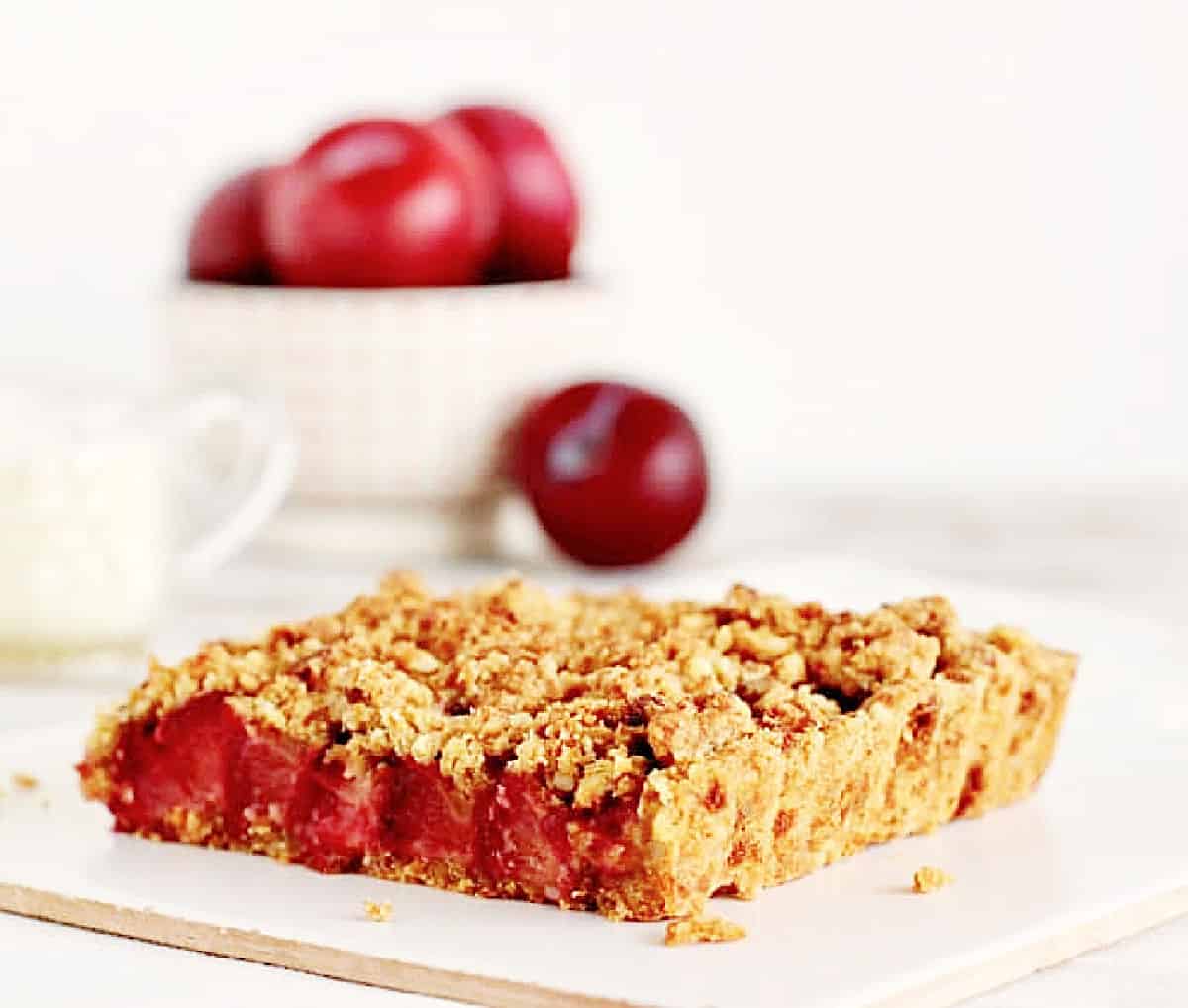 One square serving of plum crumble tart on white surface; whole plums in bowl in background