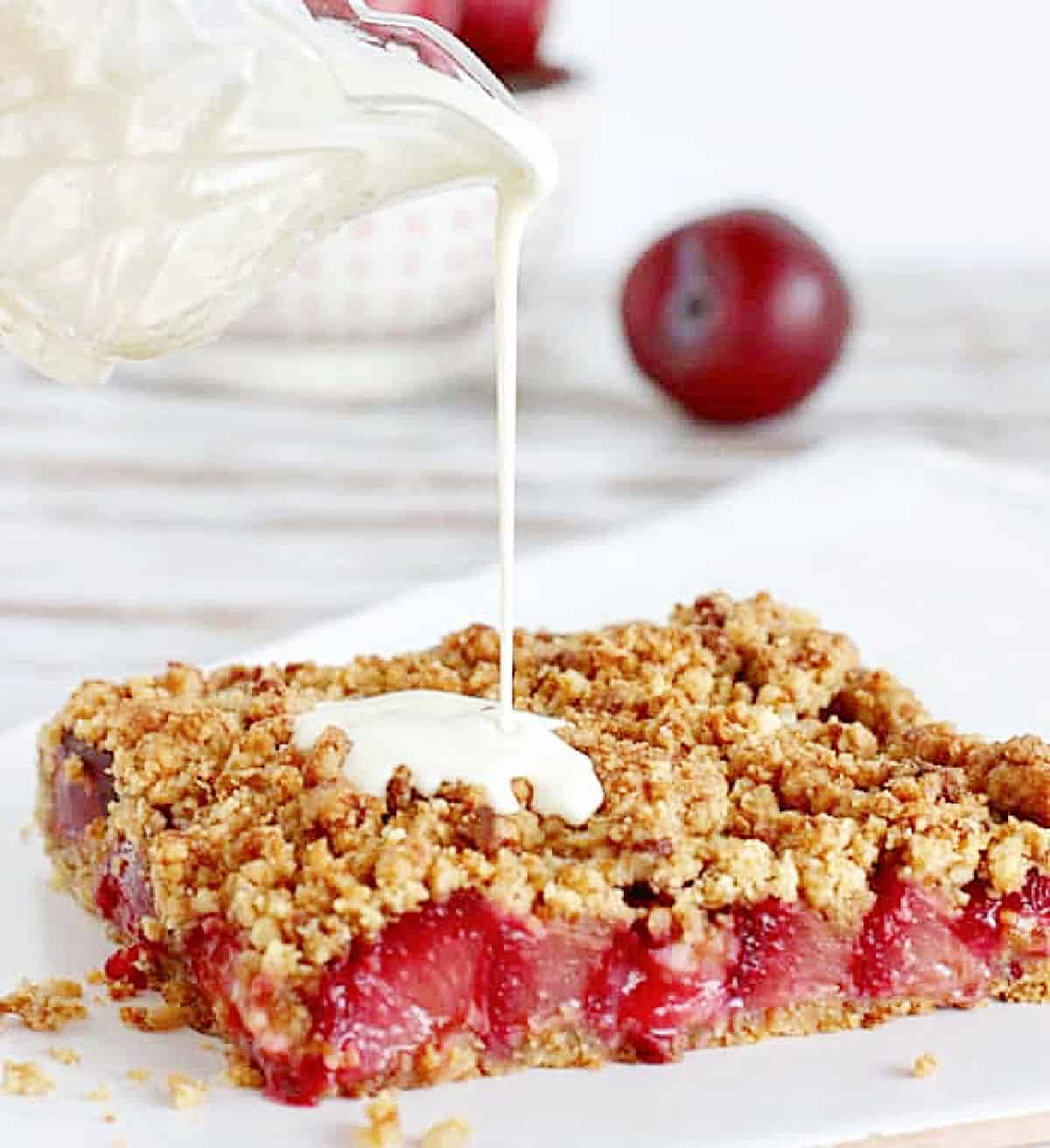 Pouring cream from jar on portion of plum crumb tart, white background with whole plum