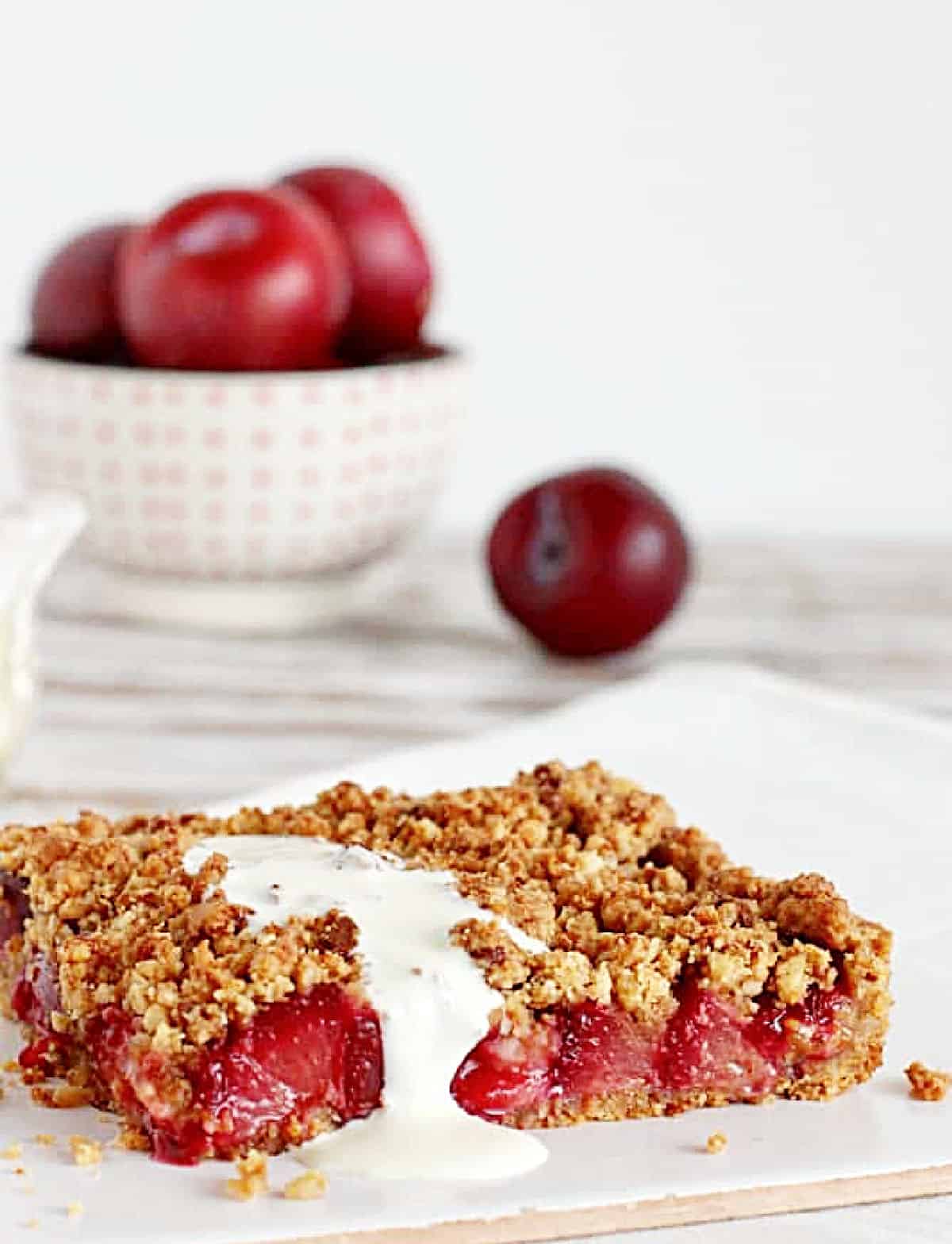 White background with fresh plums in bowl and slice of plum crumble bars with cream.