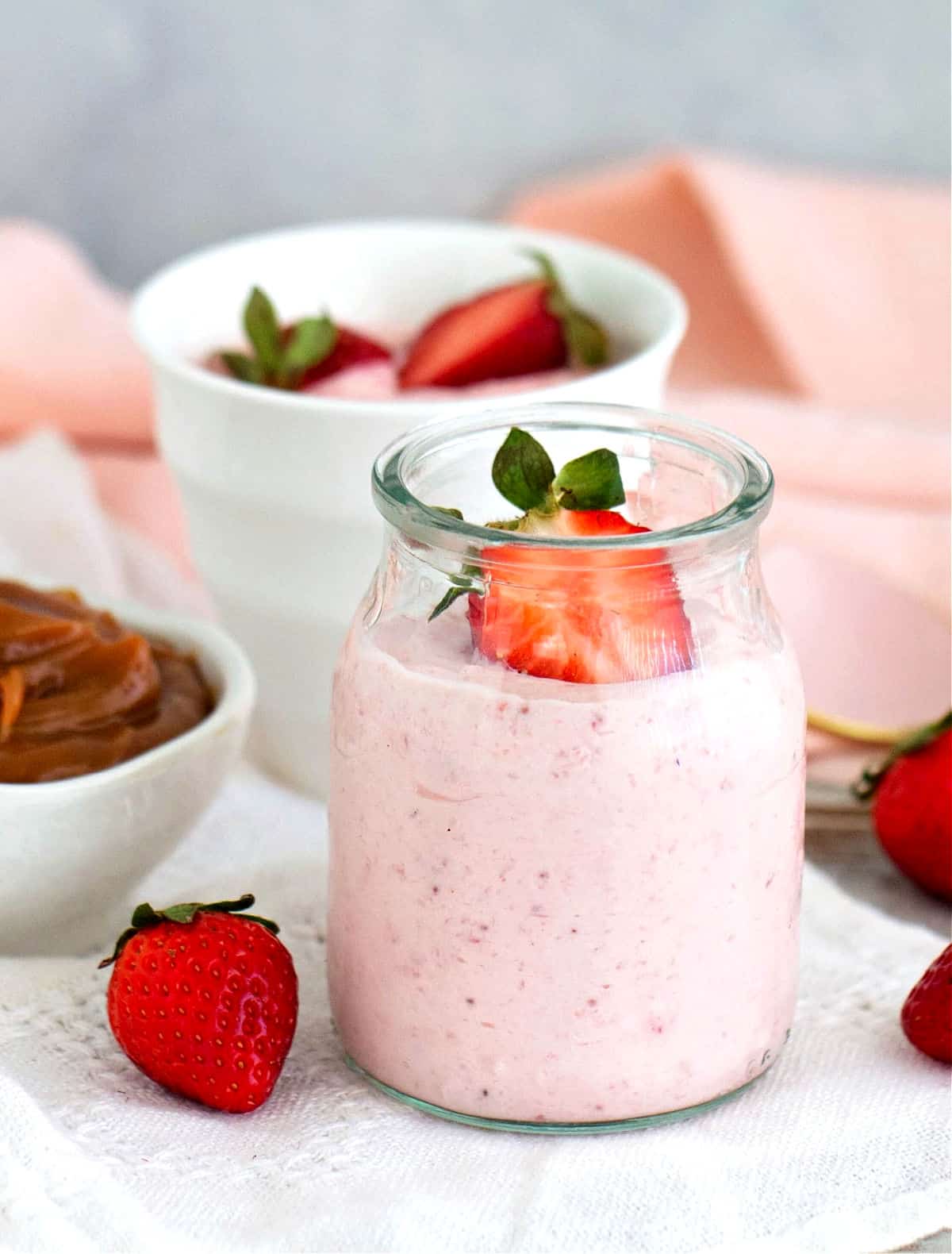 Individual serving of strawberry dessert in glass jar, bowl with berries, pink grey background.