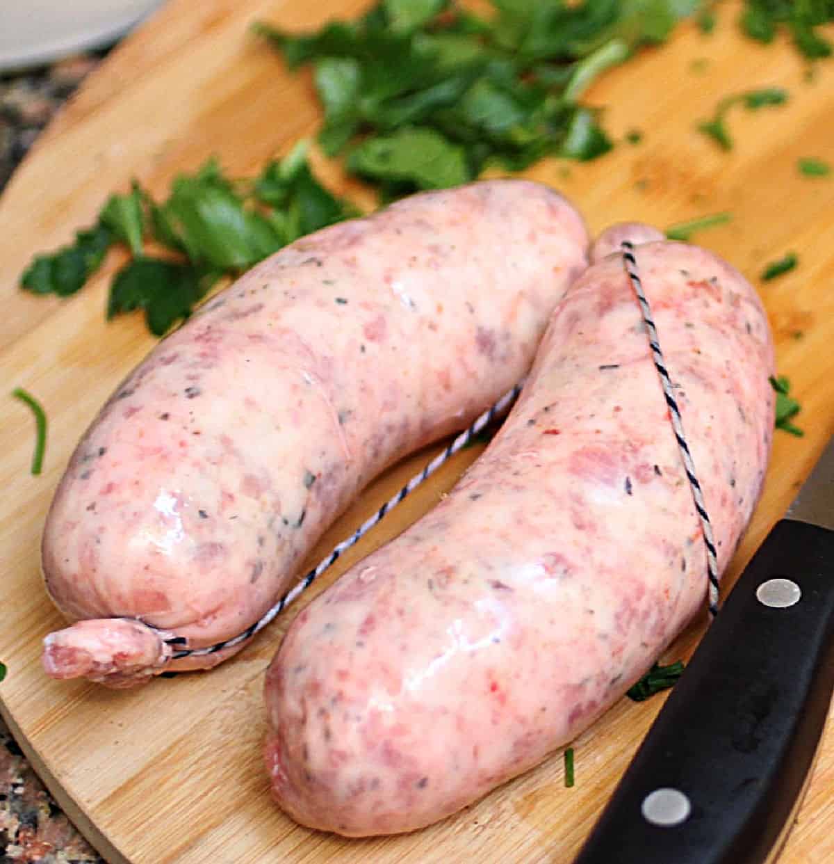 Two raw chorizos on a wooden board. Kitchen knife and chopped parsley around.