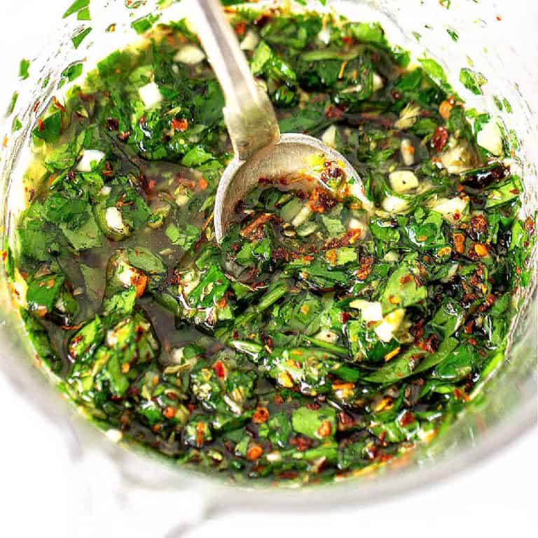 Top view of glass jar with chimichurri sauce and silver spoon, white surface