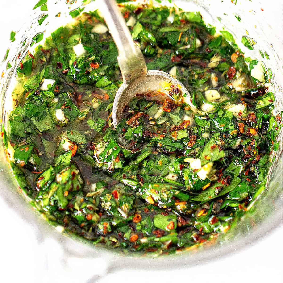 Top view of glass jar with chimichurri sauce and silver spoon, white surface.